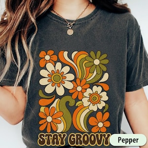 Könnte beinhalten: Dunkelgraues T-Shirt mit einem Retro-Blumenmuster in Orange, Creme und Grün. Die Worte "STAY GROOVY" sind in brauner Schrift am unteren Rand der Grafik aufgedruckt. Das Shirt hat die Farbe Pfeffer.