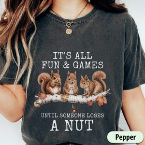 May include: Dark grey t-shirt with the text "IT'S ALL FUN & GAMES UNTIL SOMEONE LOSES A NUT." The shirt features a graphic of three squirrels on a branch. The word "Pepper" is in a green rounded rectangle.