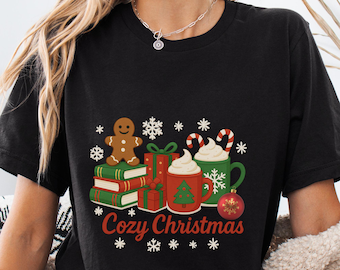 Op de afbeelding: Zwart T-shirt met een feestelijke "Cozy Christmas" print. Het ontwerp toont een peperkoekmannetje, ingepakte cadeaus, opgestapelde boeken, mokken met slagroom, zuurstokken, sneeuwvlokken en een kerstbal.