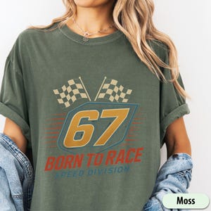 May include: Moss green t-shirt with a vintage racing graphic. The design features the number 67 in gold and blue, two checkered flags, and the text "BORN TO RACE SPEED DIVISION".