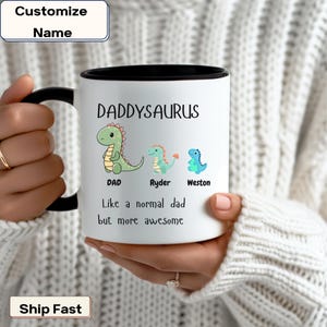 Puede incluir: Taza de café de cerámica blanca con borde y asa negros. La taza tiene una ilustración de dinosaurio verde y el texto "DADDYSAURUS" en negro. El texto "DAD" está debajo del dinosaurio, y los textos "Ryder" y "Weston" están debajo de dos dinosaurios más pequeños. El texto "Like a normal dad but more awesome" está en la parte inferior de la taza.