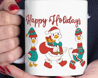 Op de afbeelding: Witte keramische mok met de tekst "Happy Holidays" in rode letters. De mok toont drie cartoon ganzen gekleed in vakantiekleding, inclusief hoeden, sjaals en kerstman outfits. Gemaakt in de VS.