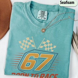 May include: Seafoam green t-shirt with a racing-themed graphic. The design features the number "67" in gold, two checkered flags, and the text "Born to Race". A silver necklace with a pendant rests on the shirt.