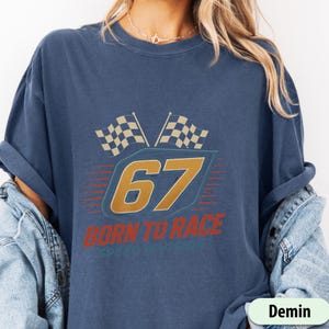 May include: A navy blue t-shirt featuring a racing-themed design. The design includes the number "67" in gold, two checkered flags, and the text "BORN TO RACE". The shirt has a vintage, worn look.