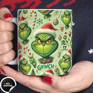 Op de afbeelding: Een witte keramische mok met een herhalend patroon van de Grinch met een kerstmuts, zuurstokken en kerstversieringen op een lichtgroene achtergrond. Het woord "Grinch" is groen gedrukt. De mok wordt door een persoon vastgehouden.
