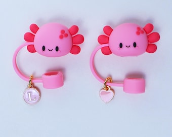 May include: Two pink axolotl-shaped straw toppers with red gills and smiling faces. Each topper has a pink loop and a charm: one with the letter "L" and the other with a heart. The toppers are designed to fit on a straw.