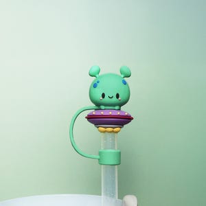 May include: A teal alien-shaped bottle stopper with a smiling face and blue spots sits atop a purple and red disc. The stopper is attached to a clear straw with a green loop. The background is a solid light green.
