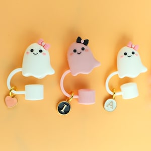 May include: Three straw covers featuring ghost figures. Two white ghosts wear pink bows, and one pink ghost wears a black bow. Each ghost is attached to a white or pink ring with a gold-toned charm, including a pink heart and circular tags with letters I and A.