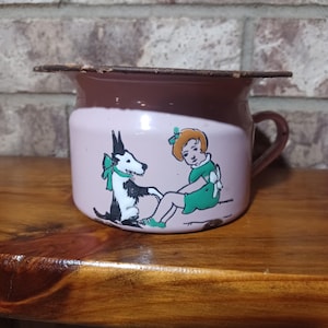 May include: Vintage pink and brown enamelware chamber pot with a handle. The pot features a cartoon illustration of a girl in a green dress and a black and white dog with a green bow. The rim has a brown trim.