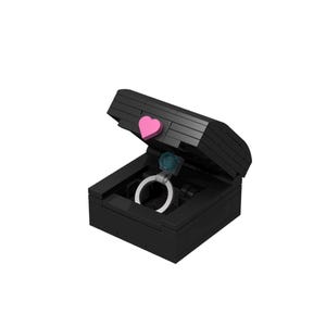 May include: A black ring box with a pink heart on the lid. The box is open and a silver ring with a blue gemstone is inside.