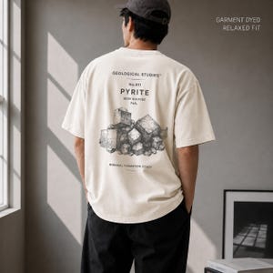 Könnte beinhalten: Ein cremefarbenes Kurzarm-T-Shirt mit lockerer Passform und stückgefärbtem Finish. Auf der Rückseite befindet sich eine schwarze Grafik aus kubischen Pyritkristallen mit dem Text Geological Studies, No.011, Pyrite, Iron Sulfide FeS2, Mineral Formation Study.