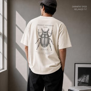 Pode incluir: Uma t-shirt de manga curta em tom off-white com corte relaxado e acabamento tingido. As costas apresentam um gráfico preto detalhado de um escaravelho com o texto INSECT STUDIES No.004 BEETLE COLEOPTERA SPECIMEN ARCHIVE.