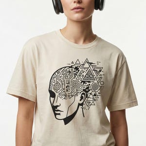 May include: A cream-coloured t-shirt with a black graphic of a human head. The left side of the head has a circuit board design, and the right side features geometric shapes. The person in the image is wearing black headphones.