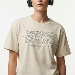 May include: A cream-colored t-shirt with a black graphic design and the text "NEURODIVERGENT FOCUS." The design features a wavy line with geometric shapes. The person in the image is wearing black headphones.