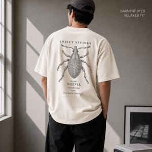 Op de afbeelding: Een off-white t-shirt met korte mouwen, een relaxte pasvorm en een garment-dyed afwerking. De achterkant toont een gedetailleerde zwarte afbeelding van een snuitkever met de tekst INSECT STUDIES, No.007 WEEVIL, Curculionidae en SPECIMEN ARCHIVE.