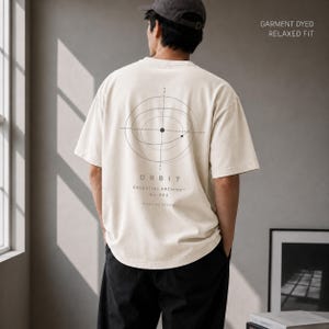 May include: A cream-colored short-sleeve t-shirt with a relaxed fit and garment-dyed finish. The back features a black graphic of orbital paths and the text "ORBIT CELESTIAL ARCHIVE No.002". The top right corner includes the text "GARMENT DYED RELAXED FIT".