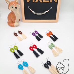May include: Assortment of handmade tassel earrings in various colors, including green, red, blue, purple, and orange. The earrings feature a black tassel and a cream-colored tassel. The earrings are displayed on a white surface with a sign that reads "Vixen Handmade".