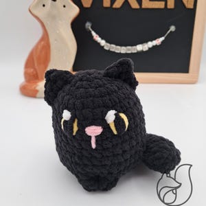 May include: Black crocheted cat toy with yellow eyes and a pink nose. The toy is sitting on a white surface. Vixen Handmade is written on the bottom of the image.