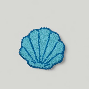 Puede incluir: Una alfombra de crochet en forma de concha, de color azul y turquesa. La alfombra es texturizada.