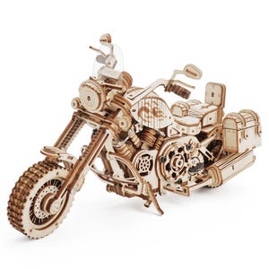 May include: A mechanical model of a cruiser motorcycle constructed from light-colored laser-cut wood. The model features intricate gear details, a front headlight, handlebars with mirrors, a fuel tank with flame graphics, a seat, and rear storage.