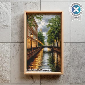 Könnte beinhalten: Eine gerahmte Kreuzstichstickerei, die eine Kanalszene in der Abenddämmerung mit einem Steingebäude, einer Brücke und grünen Bäumen zeigt. Das Wasser reflektiert den warmen gelben Schein der Straßenlaternen. Der Text BeeStitchUa.Etsy.com steht unten.