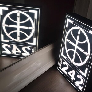 May include: Two illuminated square signs with a basketball design and the text "SAS" and "242". The signs are black with white illuminated graphics and text. The signs are on a dark brown surface.
