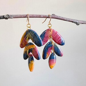 Puede incluir: Un par de pendientes colgantes con tres colgantes en forma de lágrima. Los colgantes están hechos de arcilla polimérica y presentan un diseño marmoleado en tonos azules, rosas y amarillos.