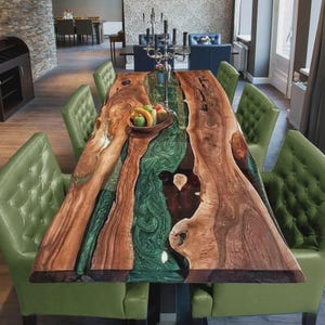 Op de afbeelding: Een grote, rechthoekige eettafel met een uniek ontwerp. Het tafelblad heeft een rivier van groene hars die tussen secties van natuurlijk hout stroomt. De tafel is omringd door groen gestoffeerde stoelen. Een fruitschaal staat op de tafel.