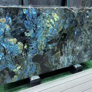 May include: Large slab of polished stone with a striking pattern of blues, greens, and blacks. The stone has a natural, textured appearance with areas of iridescent colour. The slab is supported by black blocks and rests on a green surface.