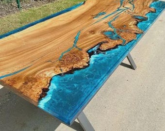 Può includere: Un tavolo rettangolare in legno con un design a bordo vivo. Il tavolo presenta un piano in legno marrone chiaro con accenti di resina blu, che ricorda un paesaggio costiero. Il tavolo è sostenuto da gambe in metallo grigio.