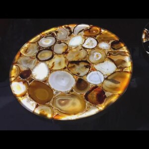 May include: A round, decorative tabletop made of polished agate stones. The stones are in shades of white, brown, and gold, with a glowing, backlit effect. The agate pieces are arranged in a mosaic pattern.