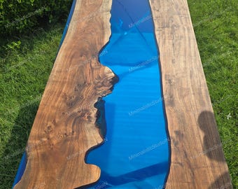Può includere: Un tavolo rettangolare in legno con un fiume di resina blu al centro. Il legno ha una venatura naturale e un colore marrone caldo. La resina blu crea un contrasto sorprendente. Il tavolo è all'aperto, su una superficie erbosa.