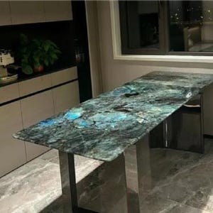 May include: A rectangular kitchen island with a polished, multi-coloured granite countertop. The granite features shades of blue, green, and grey. The island has a stainless steel base and is set against a neutral-toned kitchen backdrop.