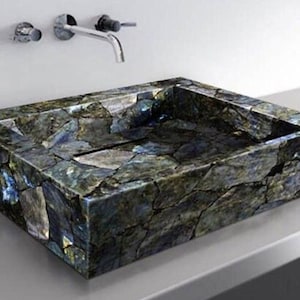 May include: A rectangular bathroom sink made of dark, multi-colored stone. The sink has a mosaic-like appearance with shades of gray, blue, and green. A chrome faucet and fixtures are mounted on the wall above the sink.