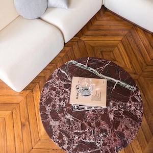 May include: A round coffee table with a red marble top featuring white veining. A stack of books and a clear glass cup sit on the table. A white sofa and wooden floor are in the background.