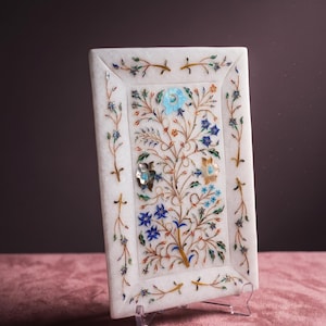 May include: A rectangular white marble tray with an inlaid floral design. The design features blue, green, and gold accents. The tray is supported by a clear plastic stand.