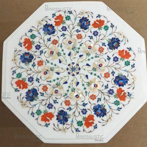 May include: White octagonal marble table with floral inlay. The design features orange, blue, and white flowers with green leaves. The table has a polished finish and is suitable for indoor use.