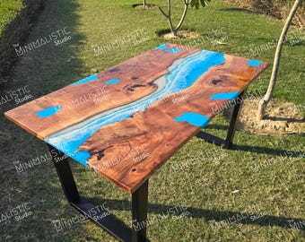 Può includere: Un tavolo da pranzo rettangolare caratterizzato da una lastra di legno naturale con bordo irregolare e un inserto centrale in resina blu brillante che ricorda un fiume. Il tavolo è sostenuto da due gambe in metallo nero a forma di U.