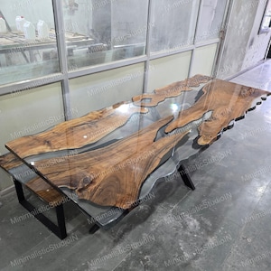 May include: A rectangular dining table featuring two natural live-edge wood slabs with a prominent grain pattern, joined by a clear, smooth resin center. The table is supported by a black metal frame with angled legs, showcasing a polished finish.
