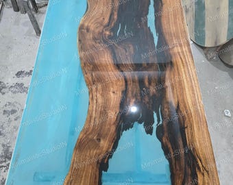 Può includere: Un tavolo rettangolare caratterizzato da una lastra di legno a bordo naturale con un marcato motivo a venature scure, integrata con una sezione in resina azzurra liscia e lucida che riempie la superficie rimanente, creando un design a fiume.