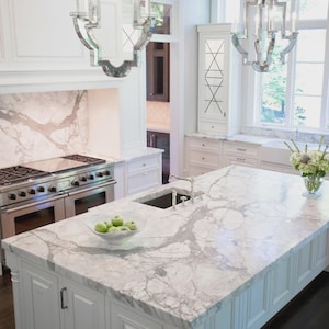 May include: A luxurious kitchen with a large marble island and countertops. Stainless steel appliances and white cabinetry complement the marble. Two ornate chandeliers hang above the island. A bowl of green apples sits on the island.