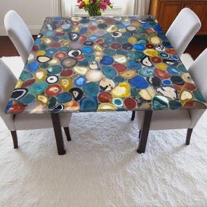 May include: A square table with a colourful agate stone top, supported by dark brown legs. Four light grey upholstered chairs surround the table, set on a white shag rug. The table's surface features a mosaic of blue, yellow, red, and white stones.
