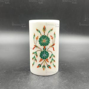 May include: A cylindrical, off-white decorative object with a floral design. The design features two green flower-like shapes with red and green leaf accents. The object is likely made of stone or a similar material.