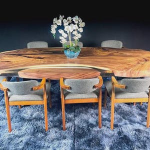 Op de afbeelding: Een grote, ovale houten eettafel met een natuurlijke rand, omringd door acht grijs gestoffeerde stoelen met houten frames. Een vaas met witte bloemen staat in het midden van de tafel. De tafel staat op een blauw tapijt.