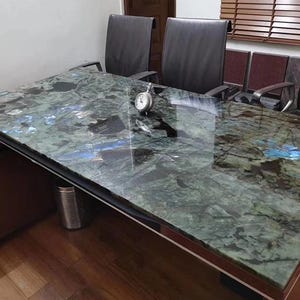 May include: A large, rectangular office desk with a polished, green and blue marble top. The desk has a dark wood frame and is surrounded by black leather chairs. A small, silver clock sits on the desk surface.