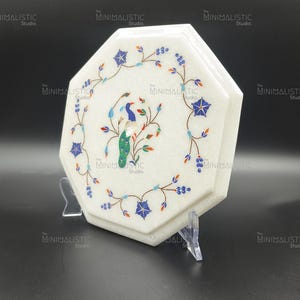May include: A white, octagonal marble box with a floral and bird inlay design. The box features blue, red, and green accents. The box is displayed on a clear stand against a black background. The text "The MINIMALISTIC Studio" is visible.