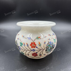 May include: A small, off-white marble vase with a wide opening and a rounded body. The vase is decorated with a floral pattern featuring red, blue, and green inlaid stones. The text "The Minimalistic Studio" is repeated around the vase.