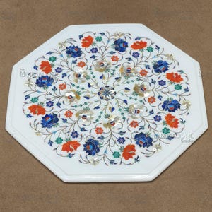 May include: White octagonal marble table with a floral inlay design. The inlay features blue, orange, and green flowers with silver accents. The table has a polished finish and is suitable for indoor use.
