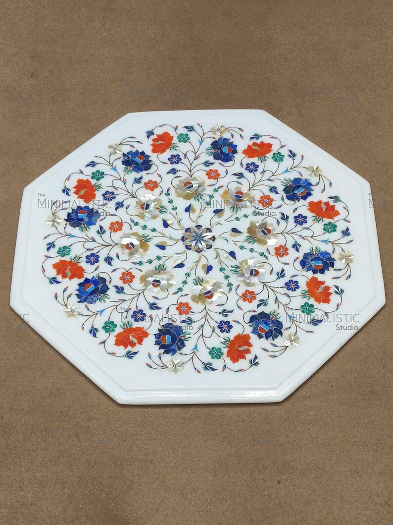 May include: White octagonal marble table with a floral inlay design. The inlay features blue, orange, and green flowers with silver accents. The table has a polished finish and is suitable for indoor use.