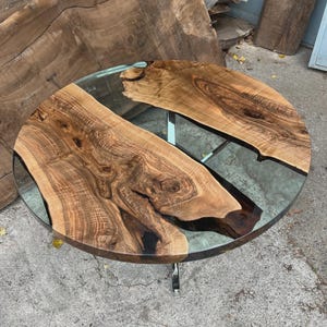 Könnte beinhalten: Ein runder Couchtisch mit einer transparenten Glasplatte und einem einzigartigen Design. Der Tisch verfügt über zwei Stücke Naturholz mit komplizierten Maserungsmustern, eingebettet in transparentem Harz. Das Gestell besteht aus verchromtem Metall.